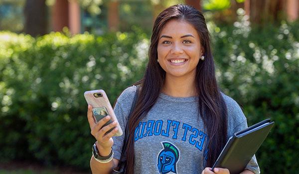 A UWF student smiles while holding books and a phone.
