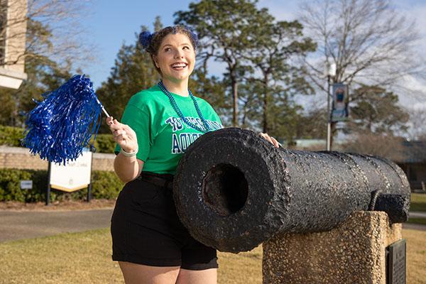 A UWF student smiles while waving a pom-pom and touching the cannon on the Cannon Green.